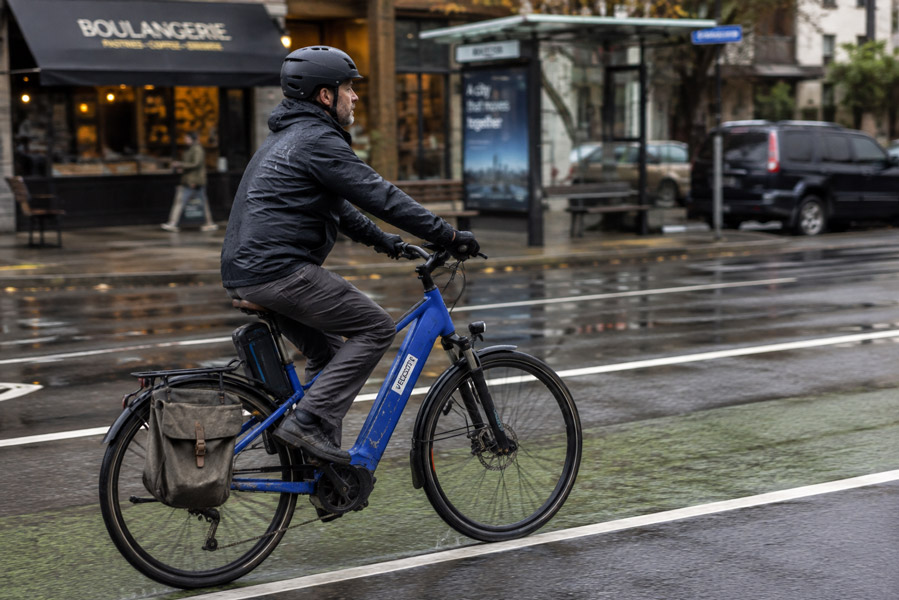 Commuter riding a blue e-bike in a city bike lane on a wet overcast morning.