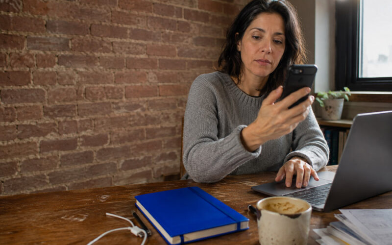 Woman at a desk checking a verification code on her phone while signing in on her laptop.