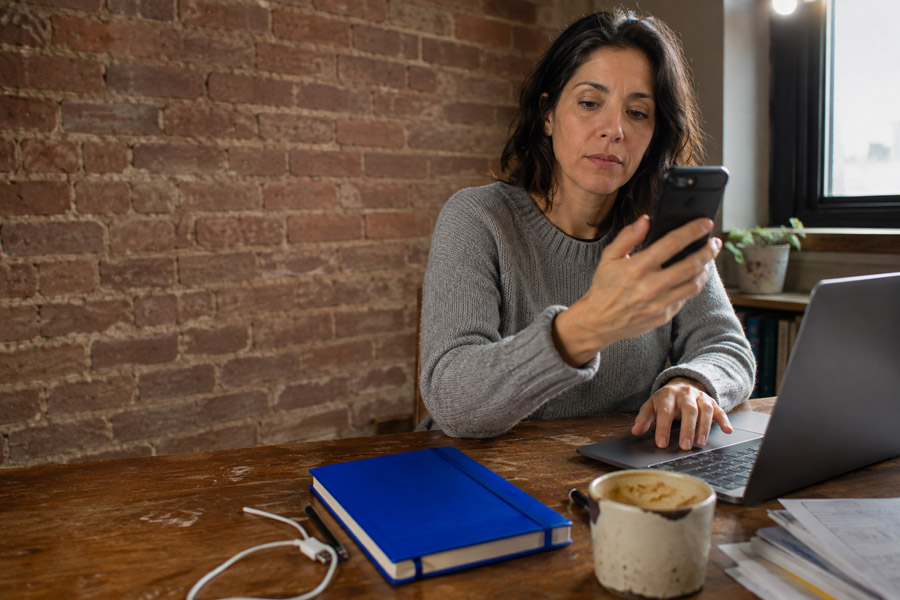 Woman at a desk checking a verification code on her phone while signing in on her laptop.